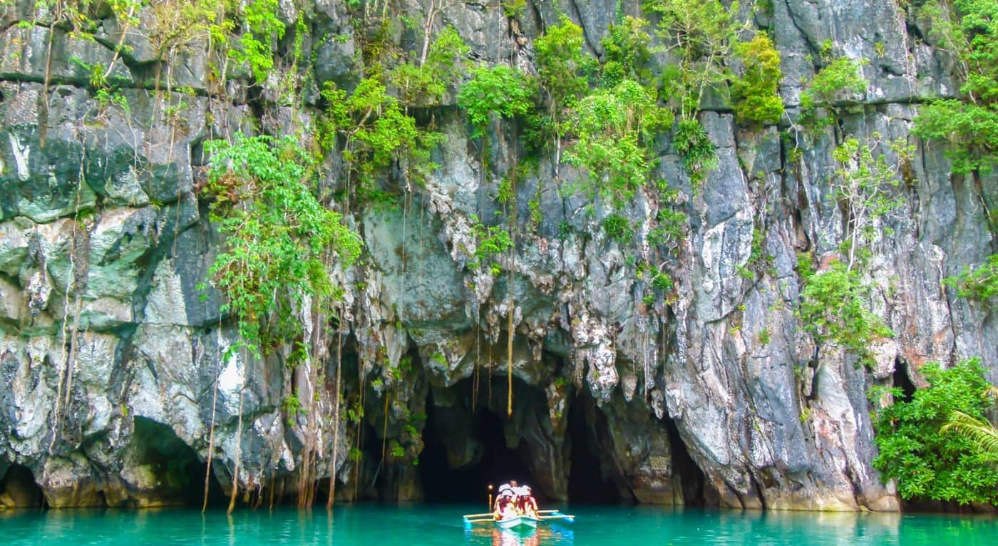 A small boat from the island-hopping tour approaches a cave entrance surrounded by lush greenery and cliffs, with turquoise water in the foreground.