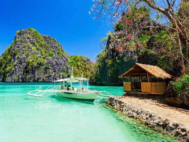 A traditional boat from the island-hopping tour floats on turquoise water near a thatched-roof hut, surrounded by lush, towering cliffs.