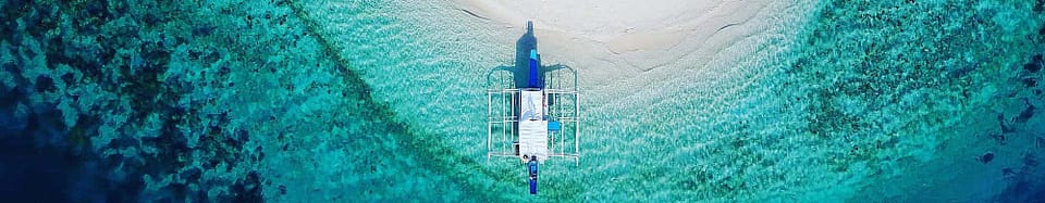 Aerial view of a small boat anchored on a clear turquoise sea near a sandy beach, perfect for island hopping among stunning coral reefs.