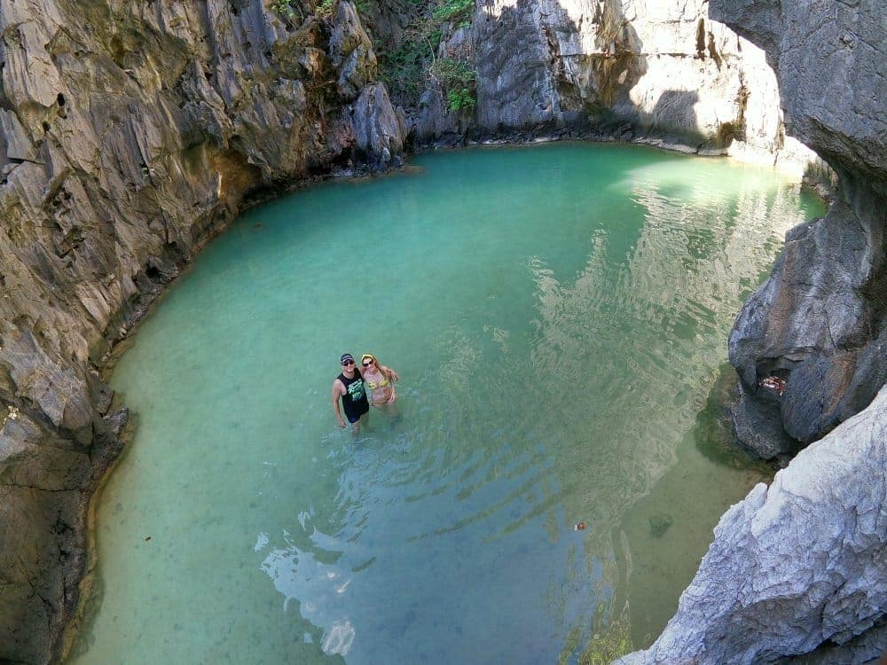 Two people stand in a clear blue pool surrounded by rocky cliffs, reminiscent of El Nido's Secret Lagoon. The calm water and sunlight illuminate the rugged textures, creating a serene setting that feels like an exclusive stop on Tour A's enchanting journey.