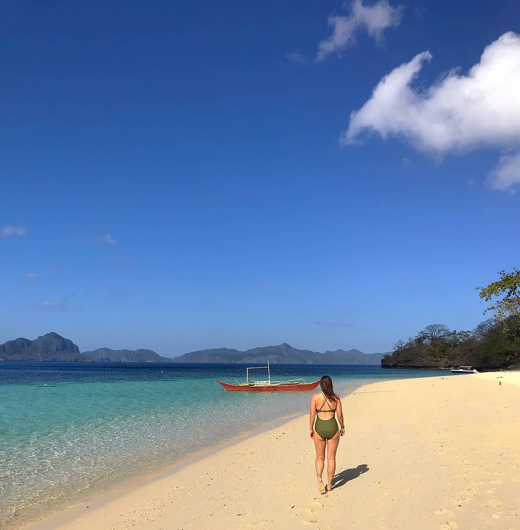 A person in a swimsuit strolls along the sandy Seven Commando Beach in Palawan, heading toward a boat on clear turquoise waters, surrounded by distant islands under a blue sky dotted with a few clouds.