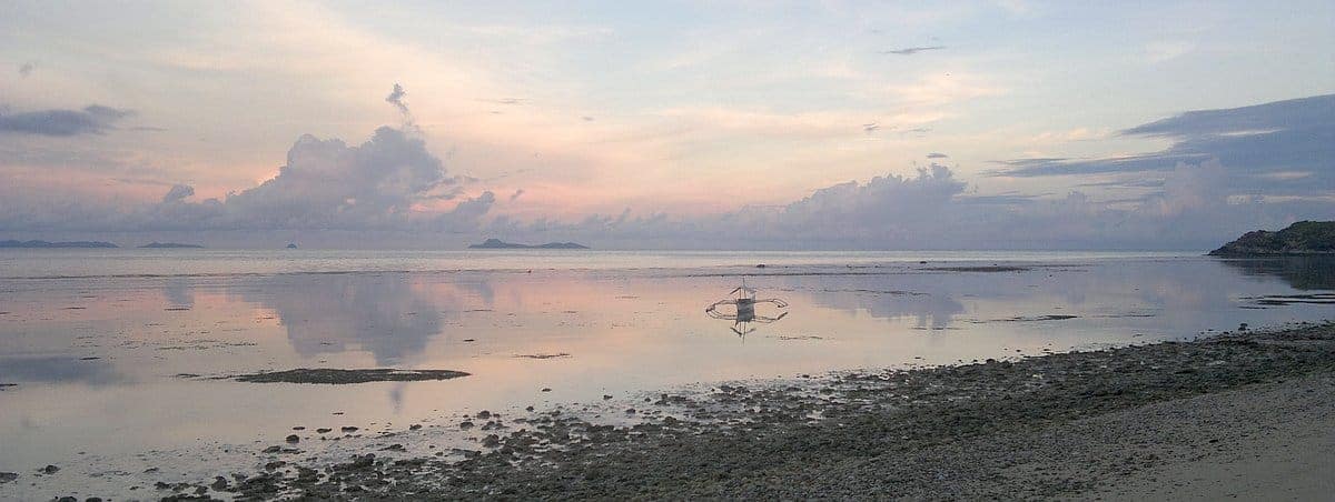 A serene beach at sunset with calm waters, where a small boat awaits its next island hopping adventure, and clouds reflect pastel colors in the sky.