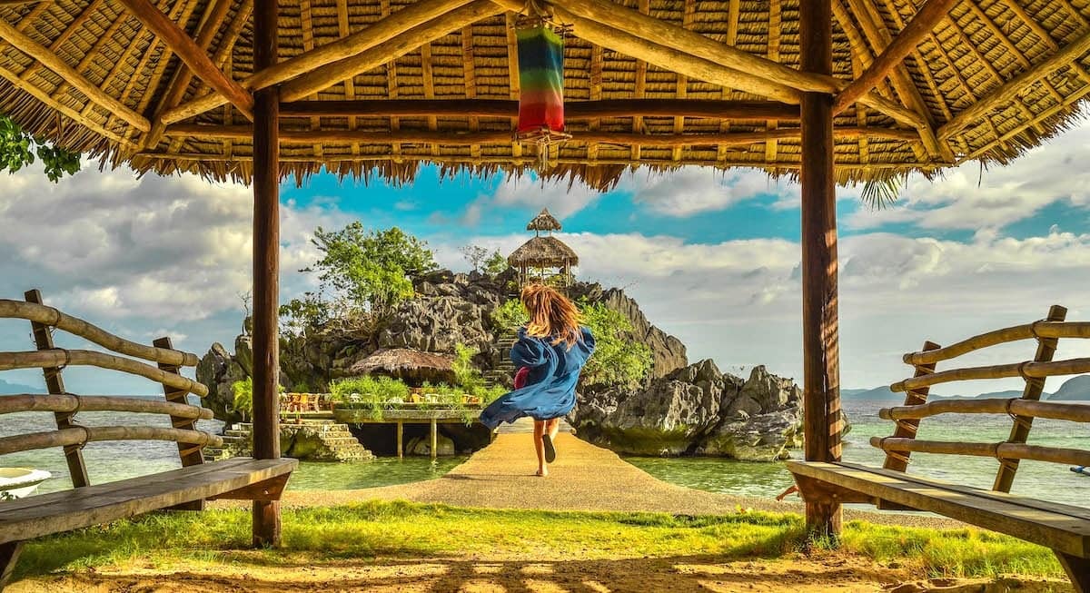 A person in a blue outfit joyfully runs on a wooden pier towards a rocky island, an ideal spot for island hopping. The scene is framed by a traditional bamboo shelter, with benches on either side, set against a bright, cloudy sky.