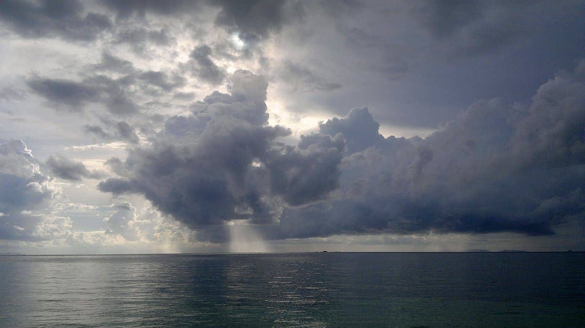 Sunrise paints the dramatic cloudy sky over a calm sea as sun rays stream through gaps in the clouds, creating a perfect backdrop for a serene boat tour.