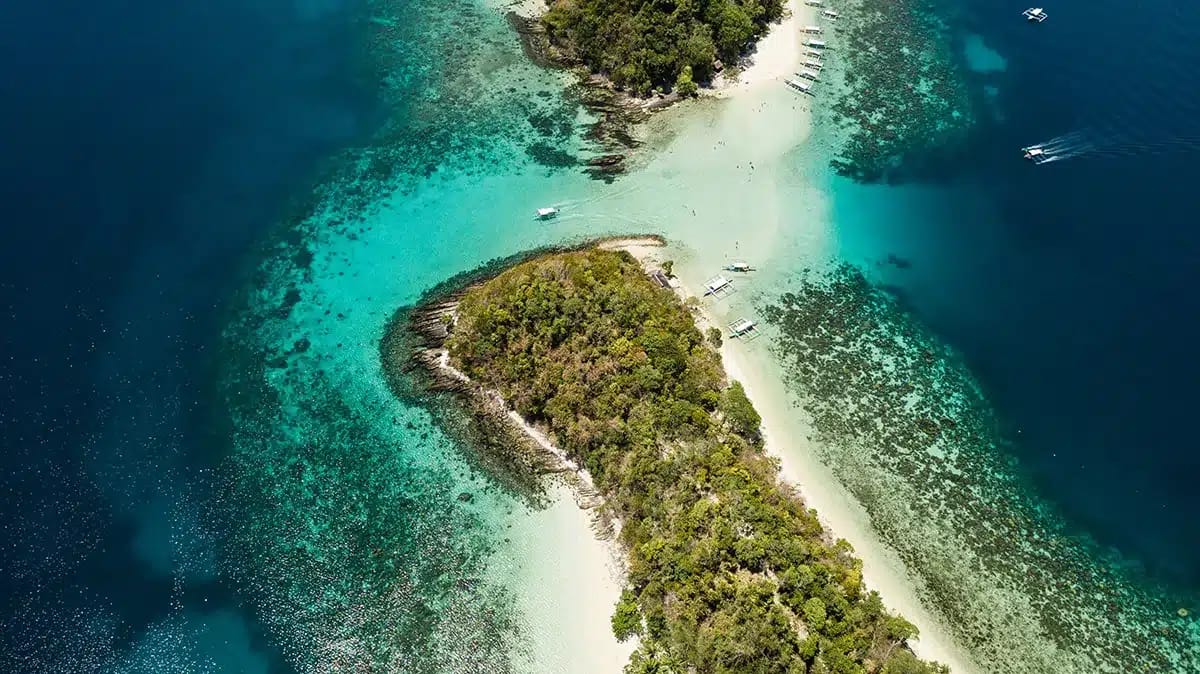 Aerial view of a tropical island, reminiscent of El Nido, with lush green vegetation surrounded by turquoise waters. White sandy beaches line the island, and a few boats are anchored nearby. Clear water reveals patches of coral reefs, making it one of the best snorkeling spots.
