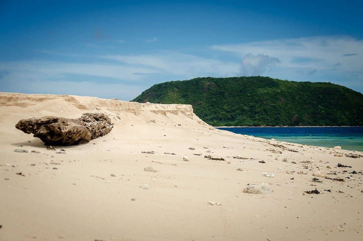 A sandy beach with scattered rocks, a distant green hill, and a clear blue sky invites you to enjoy an island hopping adventure or embark on a serene boat tour.