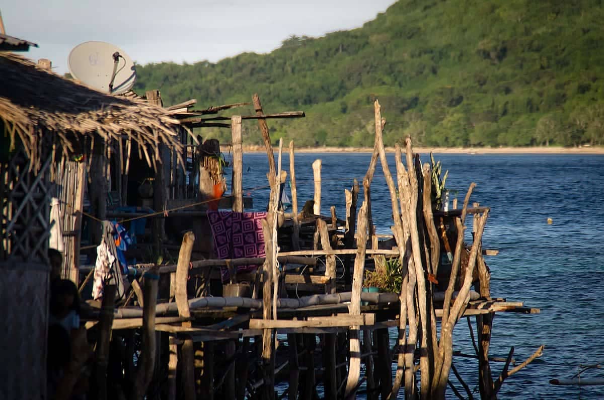 Wooden stilt houses extend over the water beside a lush green hill under a clear sky; colorful clothes hang to dry outside, offering a picturesque view perfect for those enjoying an island hopping adventure.