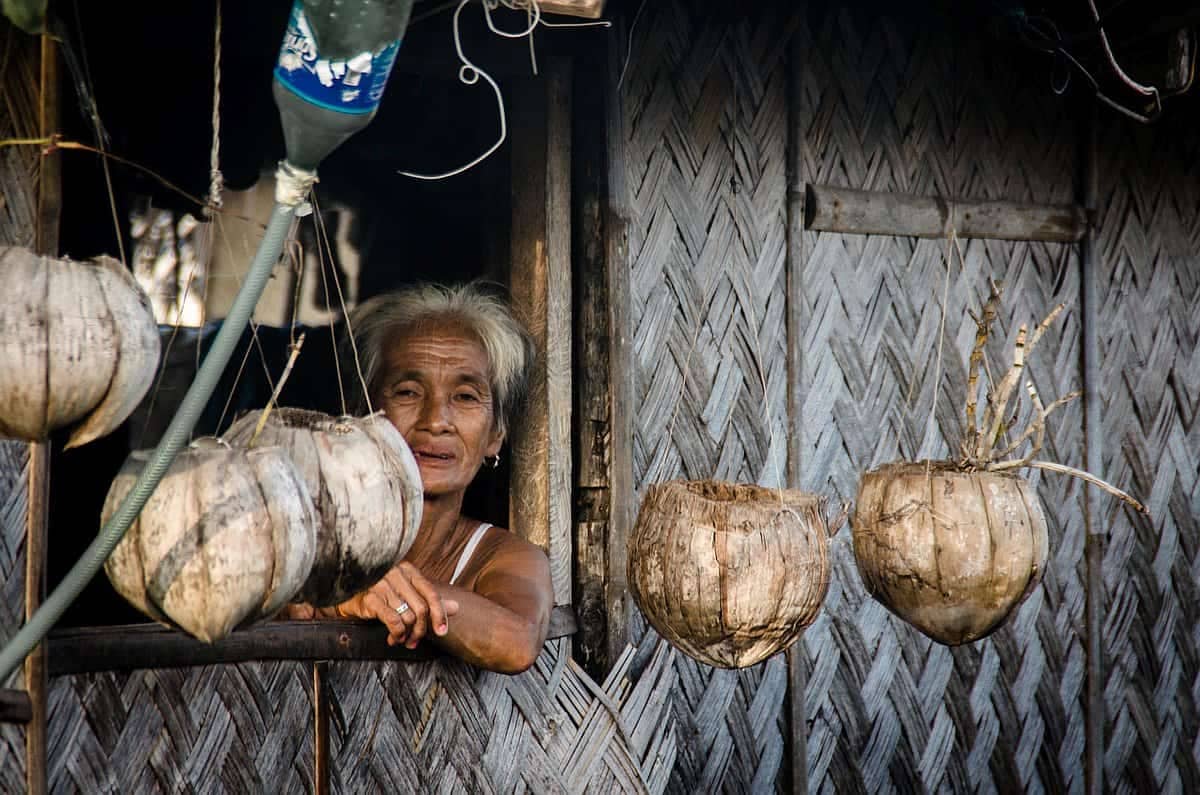 An elderly woman with gray hair stands at a window, surrounded by hanging dried coconuts, dreaming of her days spent island hopping against the backdrop of woven bamboo walls.