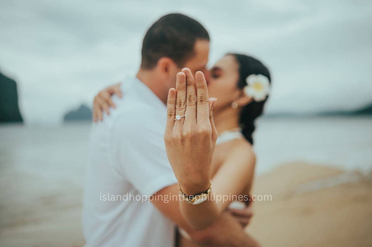 A couple embraces on a beach in Palawan, with the woman displaying a ring on her finger. A white flower graces her hair, while the blurred sand and sea evoke romance. Their blissful moment follows an enchanting island hopping adventure.