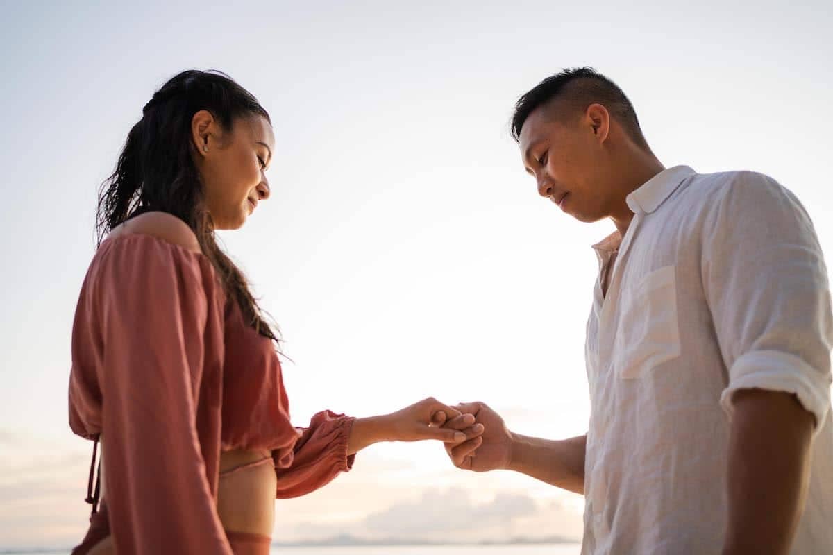 A couple stands outdoors under a clear sky, holding each other's hands gently. The woman in a pink blouse gazes down, while the man in a white shirt looks at their hands. The scene conveys a tender proposal and deep connection as they embrace the promise of marriage.