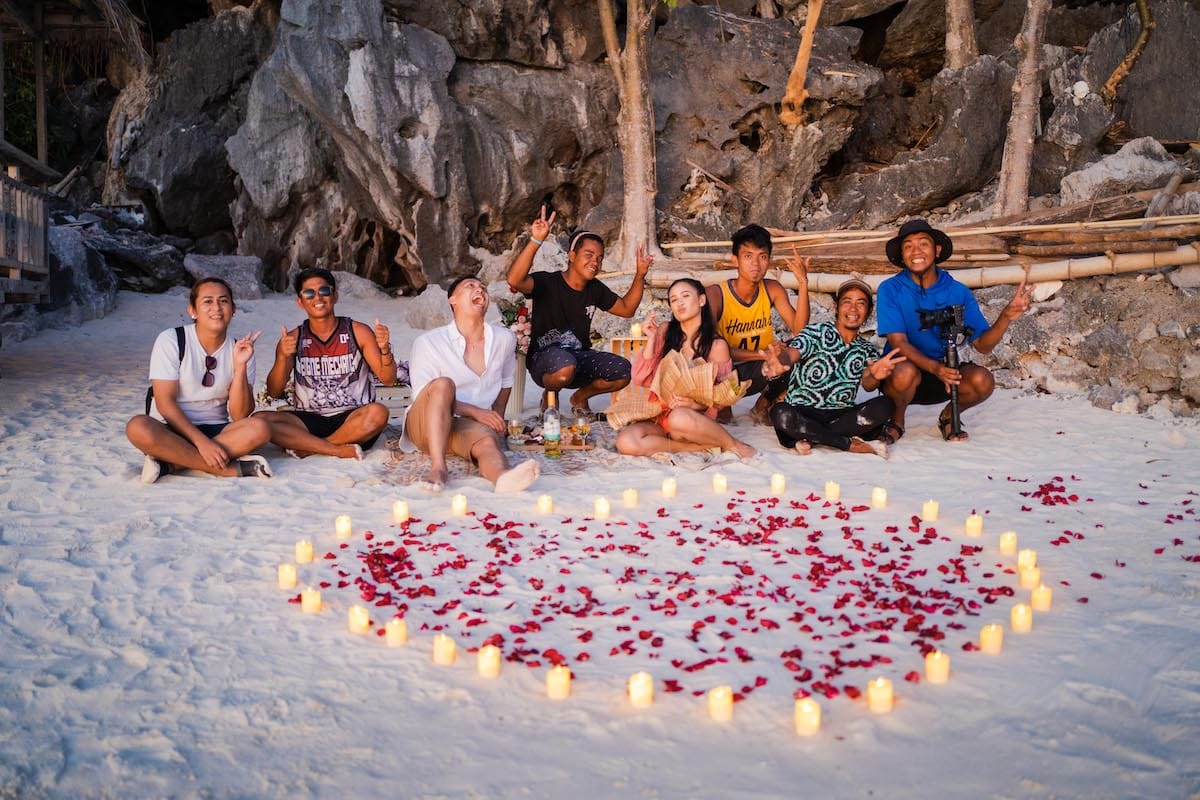 Eight friends sit joyfully on a beach, encircled by a heart of rose petals and candles, capturing the moment with rocky cliffs and trees framing their joy. It's a perfect setting for an unexpected marriage proposal amid nature's backdrop. Eight friends sit joyfully on a beach, encircled by a heart of rose petals and candles, capturing the moment with rocky cliffs and trees framing their joy. It's a perfect setting for an unexpected marriage proposal amid nature's backdrop.