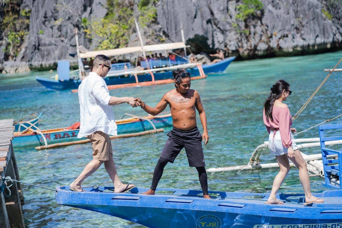 A man helps another man step onto a blue boat as a woman walks ahead, perhaps contemplating a proposal. The background reveals clear turquoise water, a wooden boat, and rugged cliffs adorned with greenery. It's a sunny day, perfect for a casual tour of this picturesque setting.