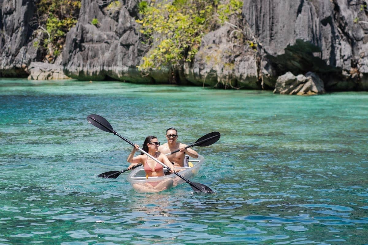 A couple enjoys a tour in a transparent kayak on clear turquoise water, surrounded by lush greenery and steep rock formations. The scene is bright and sunny, capturing a sense of adventure and tranquility—perfect for a unique marriage proposal.