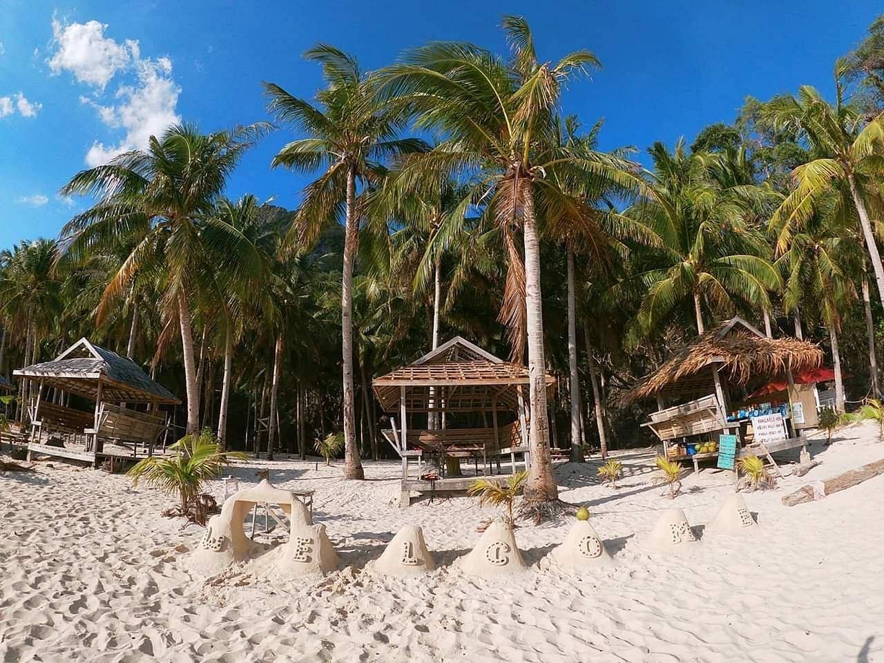 A tropical beach scene with palm trees, sandy sculptures spelling "WELCOME," and several wooden huts. The sun shines brightly in a clear blue sky at Seven Commando Beach in El Nido, Palawan.