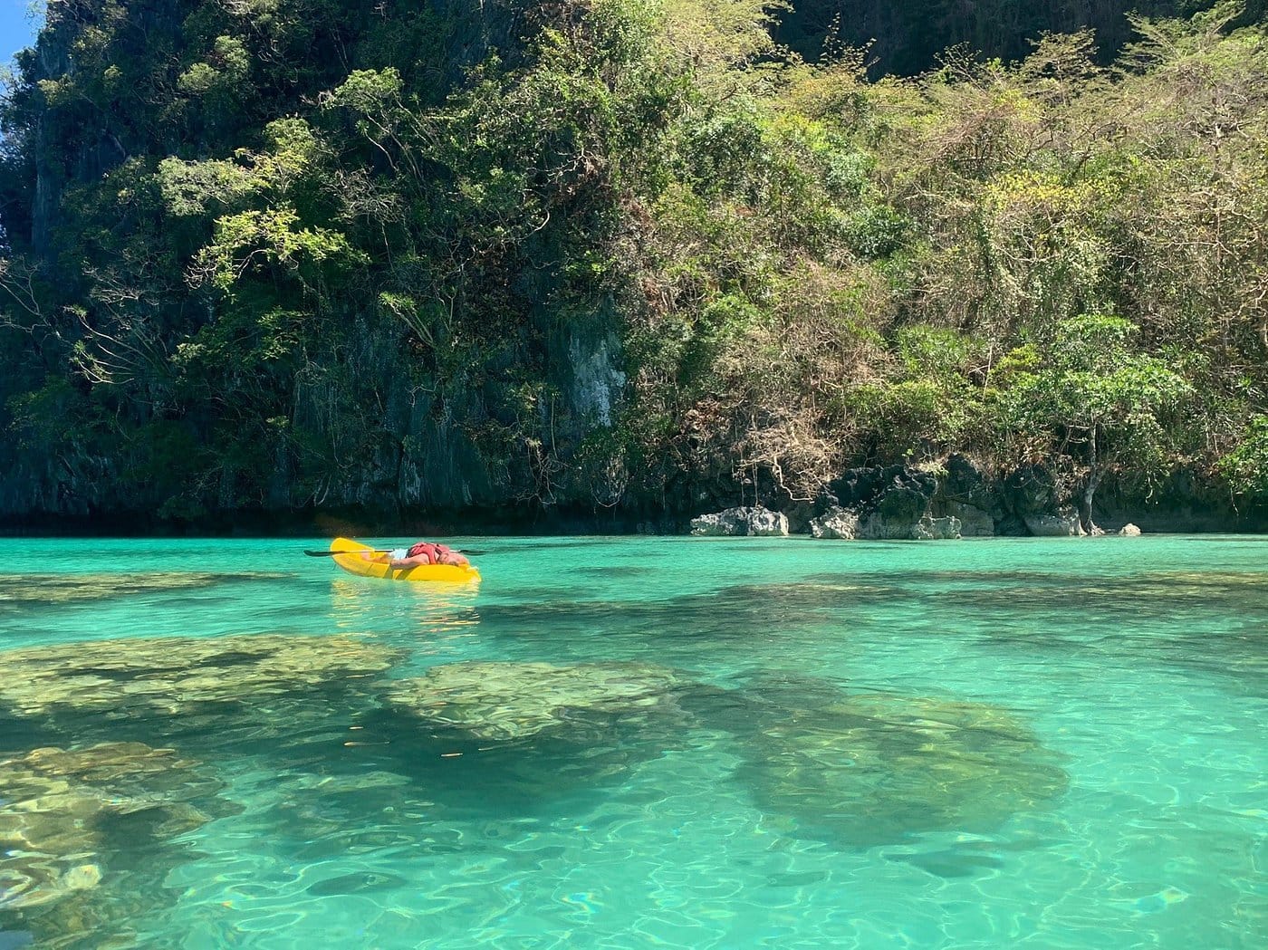 A person in a yellow kayak paddles through the clear turquoise waters of El Nido's Big Lagoon, surrounded by lush green cliffs under a sunny sky. Large rocks are visible beneath the surface, showcasing the natural beauty of Palawan.