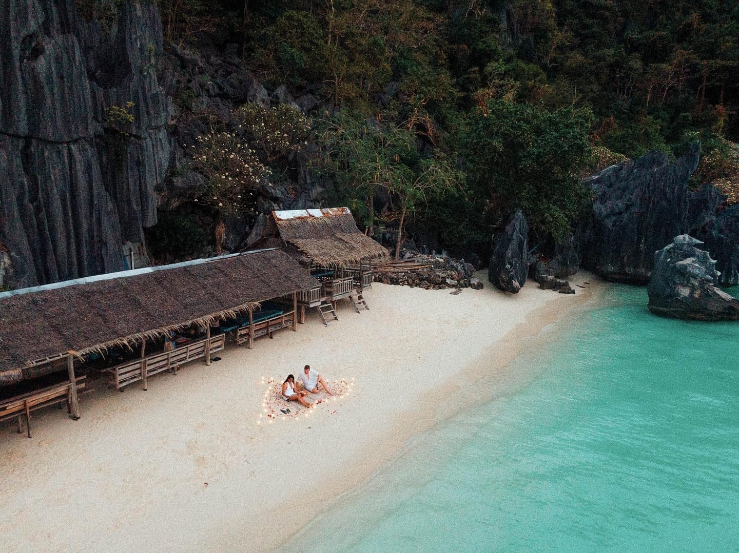 A couple sits on a sandy Banol Beach surrounded by scattered petals and candles. They are near a wooden hut with a thatched roof, close to turquoise waters. Lush greenery and rocky cliffs, ideal for island hopping adventures, envelop the serene setting.