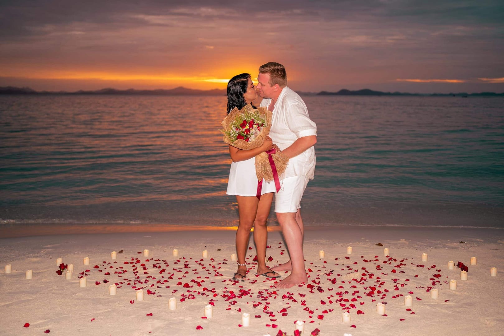 A couple kisses on a Palawan beach at sunset, surrounded by rose petals and candles arranged in a heart shape. The woman holds a bouquet of red roses. The ocean and the vibrant Coron sky paint the perfect backdrop.