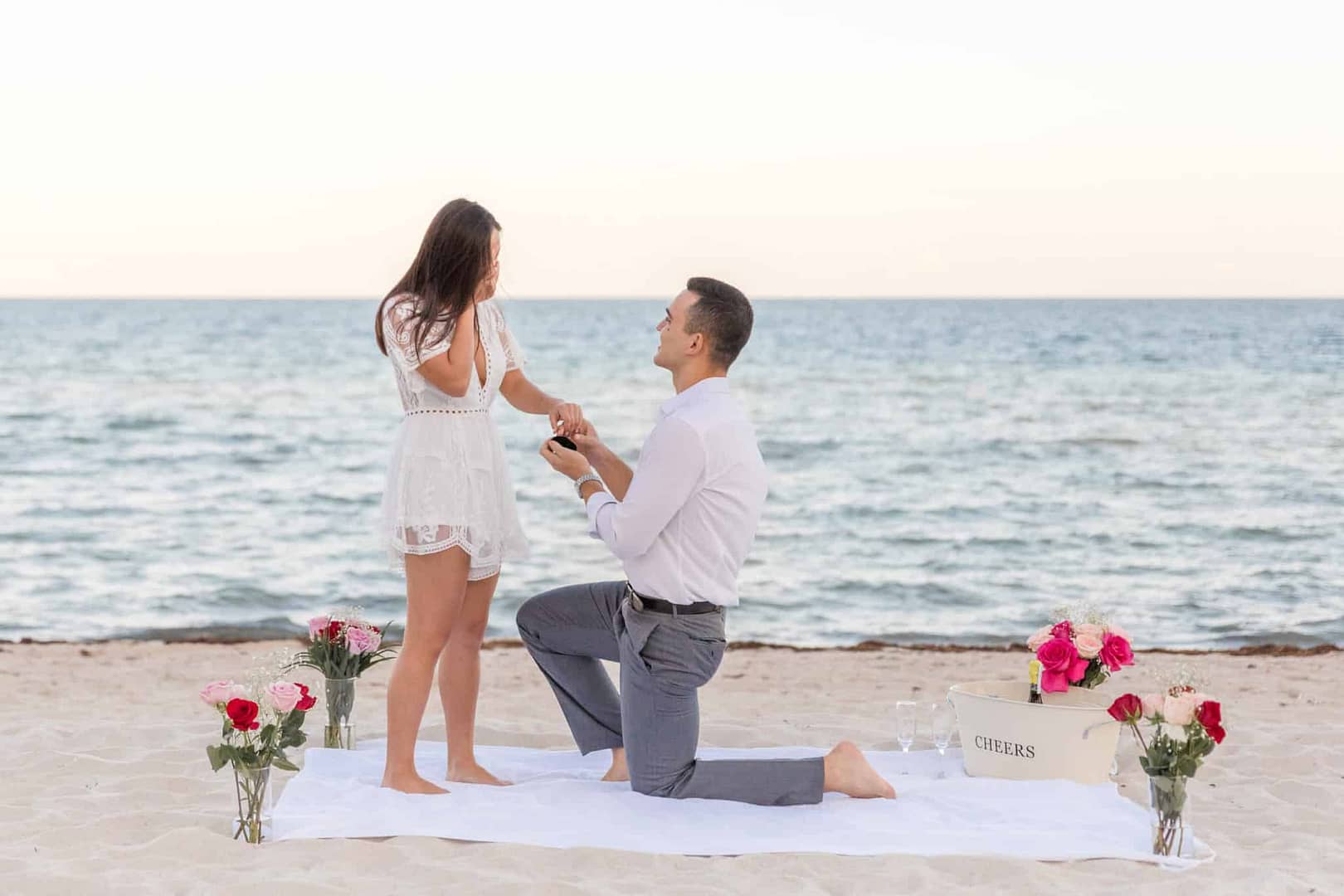 A person kneels on a beach in Palawan, proposing to another who stands with a surprised expression. They're on a white blanket surrounded by red and pink roses near the ocean. It's a romantic sunset setting, the perfect moment for an unforgettable surprise proposal during their romantic vacation.