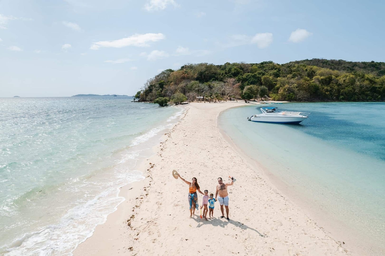 A family of four stands on a sandy beach in Palawan, waving at the camera. The beach stretches between the sea and a clear blue lagoon near Coron. A speedboat is anchored nearby, and lush Ditaytayan Island is visible under a partly cloudy sky.