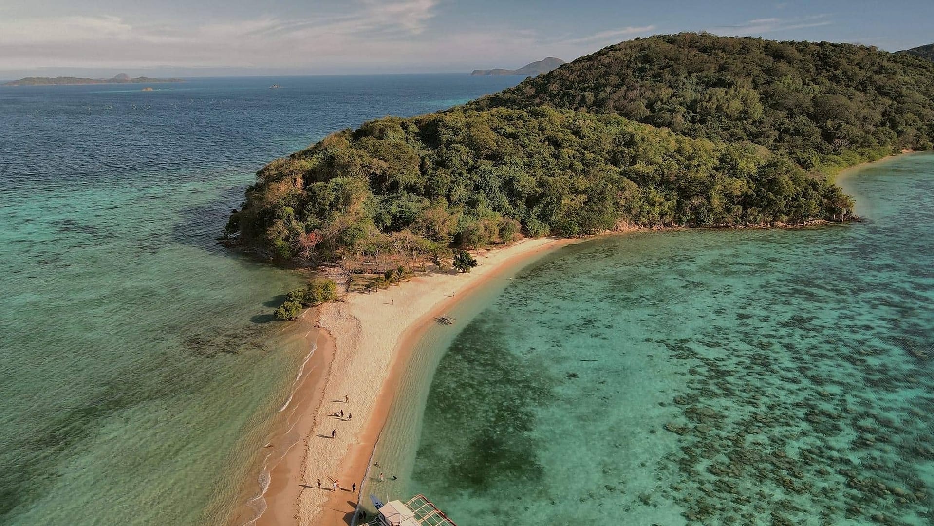 Aerial view of Ditaytayan Island in Palawan reveals a lush green hill and sandy beach embraced by clear turquoise waters. A narrow sandbar connects the island to the foreground, with a few people and a small boat near the shore, capturing the serene beauty of Coron.