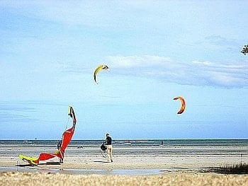 A beach scene with a person strolling on the sand near vibrant kiteboarding sails. The sky is clear with a few clouds, and the ocean stretches in the background, inviting adventurous souls to explore island hopping delights.