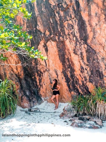 A person in swimwear leans against a large rock face on a sandy beach, with lush green plants and trees nearby, savoring a moment of relaxation after an exhilarating island-hopping boat tour.