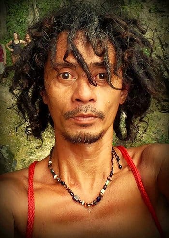 A person with curly hair and a necklace takes a selfie outdoors, capturing the excitement of an island-hopping adventure.