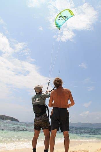 Two men on a beach prepare for kitesurfing, with one adjusting the lines in his harness and the other holding the kite bar. The clear sky and ocean set the scene, perfect for combining this thrill with island hopping to explore nearby shores.