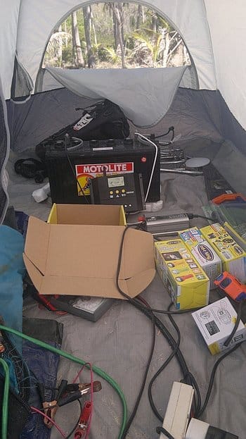 Inside a tent, with a car battery, cables, tools, and a cardboard box scattered on the floor. Forest visible through the tent entrance, reminiscent of Palawan's lush greenery experienced during island hopping adventures.