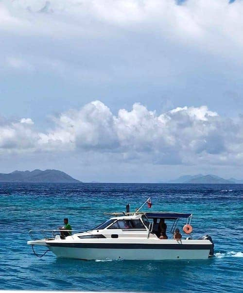 A small white motorboat with people on board is island hopping through clear blue waters. In the background, distant islands and a cloudy sky create a picturesque scene. The boat, equipped with a lifebuoy and a flag at the stern, gracefully glides along its adventure.