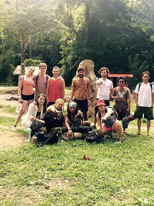 A group of people posing outdoors on grass, with trees and a large rock in the background, reminiscent of an island hopping adventure. Some are standing, while others are kneeling.