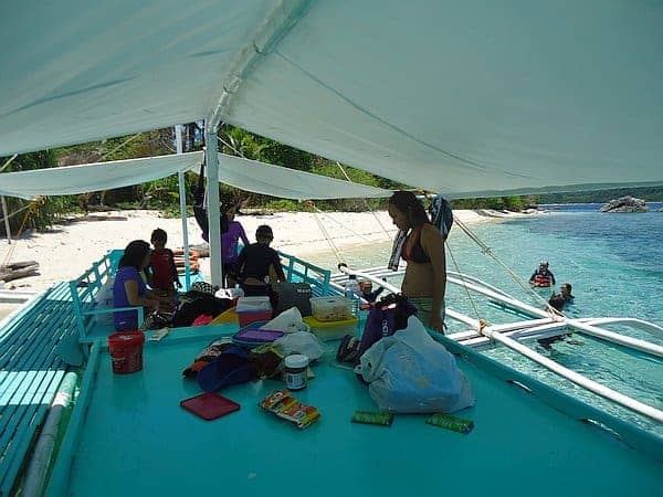 People are gathered on a boat under a canopy, docked near a sandy beach, enjoying an island hopping adventure in the Philippines. Various items like bags and snacks are on a table. The clear blue water and a few swimmers are visible, with lush green trees lining the shore in the background.