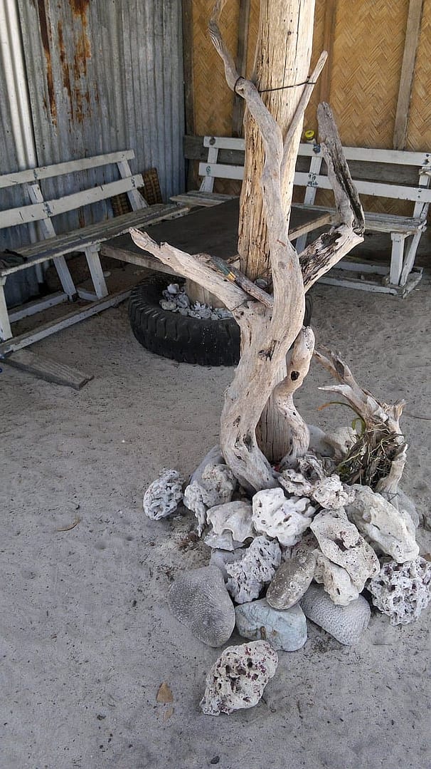 A rustic outdoor setting features a wooden pole wrapped with twisted driftwood, surrounded by a circle of coral stones. Echoing the spirit of island hopping, old wooden benches and a large black tire rest on sandy ground in the background.