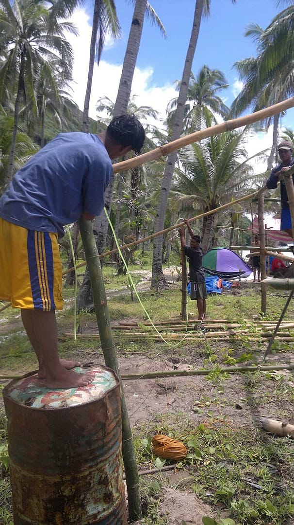 Three people working together outdoors, using bamboo poles and ropes to construct a structure among palm trees, as if preparing for island hopping. One person stands on a barrel, adjusting a pole. A tent and tropical vegetation are visible in the background.