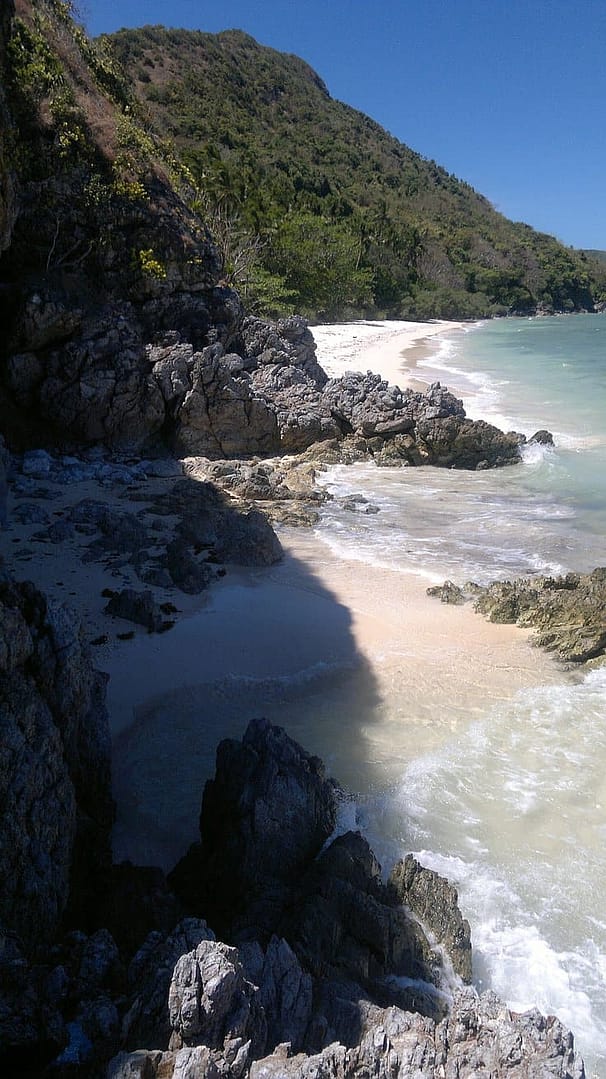 A rocky shoreline with waves gently crashing against the rocks invites adventure seekers to explore its charm. Nestled between the rocks is a small sandy beach, perfect for a pause during island hopping, with lush green hills rising in the background under a clear blue sky.