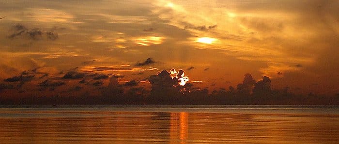 A golden sunset over the ocean casts orange and yellow hues on the water, with clouds silhouetted against the sky, creating a perfect backdrop for an enchanting boat tour while island hopping near Palawan.