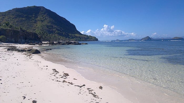 A serene beach with clear turquoise water, a rocky shoreline, and a lush green hill under a clear blue sky awaits you in Palawan—perfect for island hopping adventures.