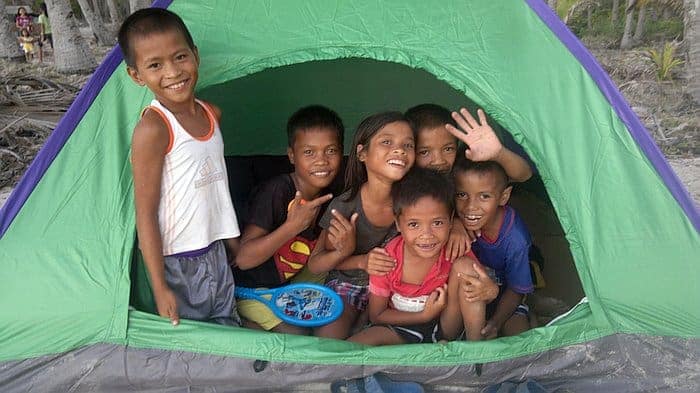 A group of smiling children in a green tent, with one child holding a toy, enjoy a sunny day dreaming of island hopping adventures and exciting boat tours in Palawan.