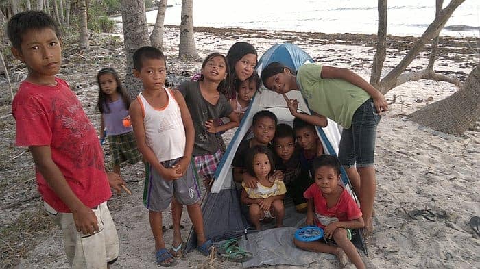 A group of children gathered around a small tent on a sandy beach in Palawan, with trees and shoreline in the background, as they excitedly discuss their next island hopping adventure.