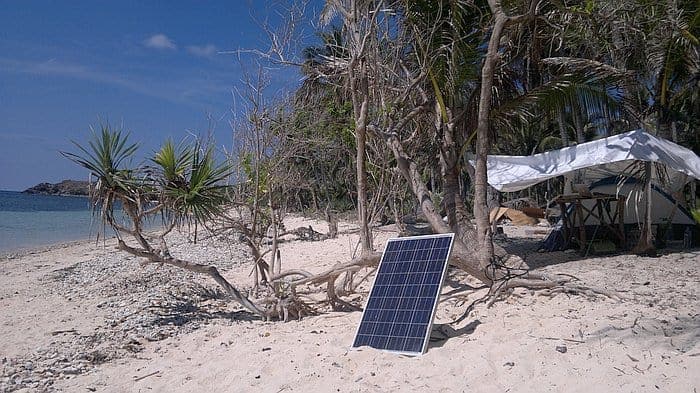 A solar panel on a sandy Palawan beach with trees, a tent, and the ocean in the background under a clear blue sky invites dreams of island hopping adventures.