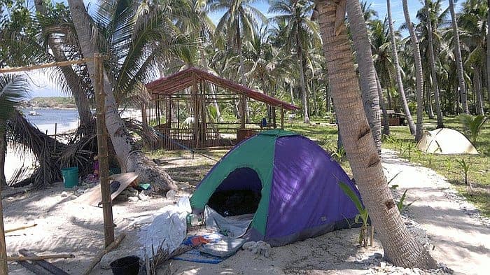 A purple and green tent is set up on a sandy beach surrounded by palm trees, capturing the essence of island hopping in Palawan. In the background, there's a small wooden structure with a red roof. A glimpse of the ocean and clear blue sky can be seen as another tent is visible in the distance.