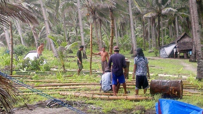 People assembling bamboo structures in a tropical setting, surrounded by palm trees and huts, prepare for an enchanting boat tour that promises to reveal the hidden beauty of the lush landscape.