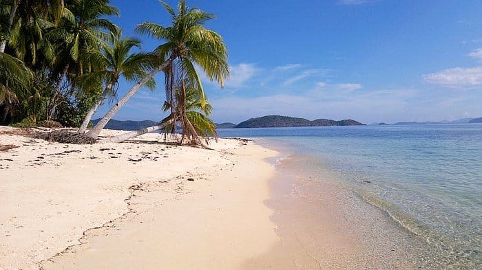 Tropical beach with clear blue water, white sand, and leaning palm trees under a sunny sky. Distant islands are visible on the horizon, inviting you to consider a relaxing boat trip.