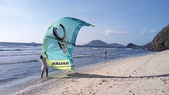 A person stands on a sandy beach, holding a large teal kite, while gentle waves lap at the shore. Distant mountains and clear blue skies invite thoughts of island hopping, as boat tours offer new horizons.