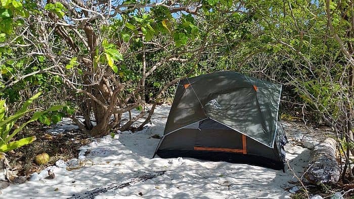 A green and gray tent is set up on sandy ground surrounded by the lush, leafy vegetation of Dimancal Island. Sunlight filters through the branches, casting dappled shadows on the tent and sand, while a coconut rests nearby under a tree—a perfect camping spot after thrilling boat tours.