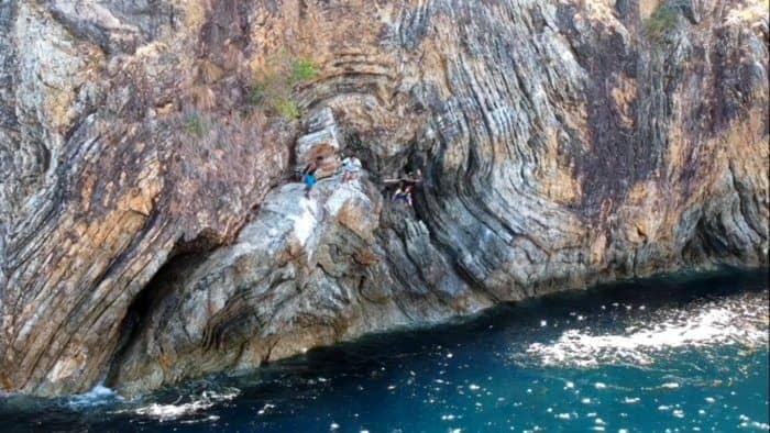 People sitting on a rugged, rocky cliff face near the blue ocean of Palawan. The rock has distinct swirling patterns and colors, and sunlight reflects off the water below.