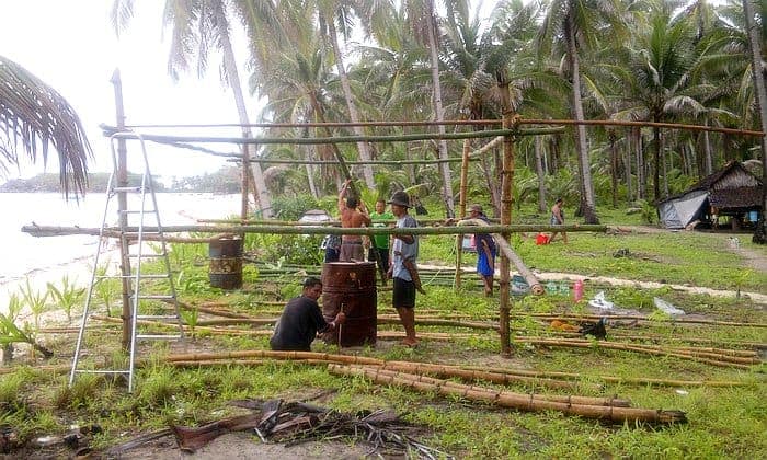 People constructing a bamboo structure on a tropical beach with palm trees in the background, preparing it as a welcoming point for an upcoming boat tour to explore the island's hidden gems.