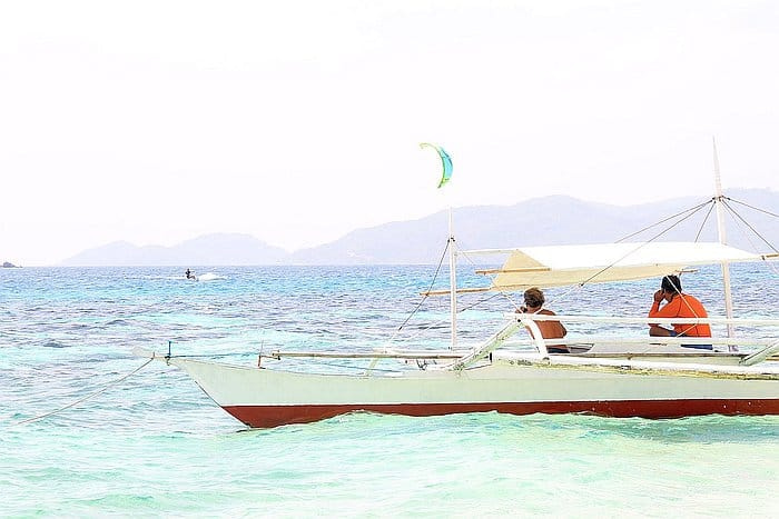 A traditional boat with two people in it floats on turquoise waters, perfect for serene boat tours. A kite surfer is visible in the background under a clear sky with distant mountains on the horizon, offering a glimpse of enticing island hopping adventures.