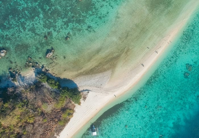 Aerial view of a narrow sandbar in Palawan, surrounded by turquoise waters. Sparse greenery is on the left, and a small boat is docked at the sandbar’s edge. Shadows of a few people walking are visible on the sand—perhaps planning an island hopping adventure or even a spontaneous marriage proposal.