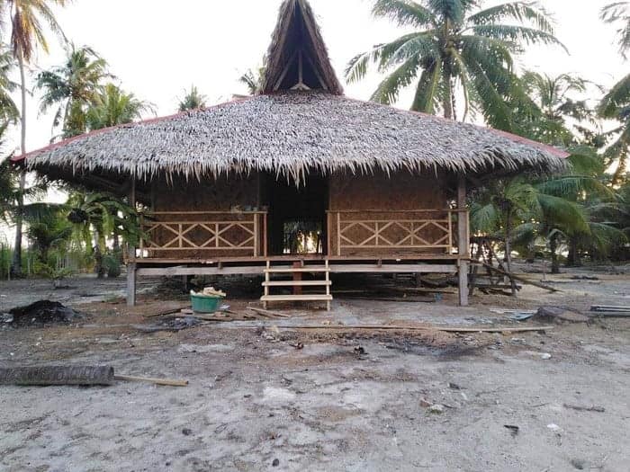 A traditional thatched-roof hut on stilts, reminiscent of those in Palawan's El Nido, is surrounded by palm trees. The hut features wooden railings and a small staircase leading up to the entrance, with boat tour materials scattered around the area.